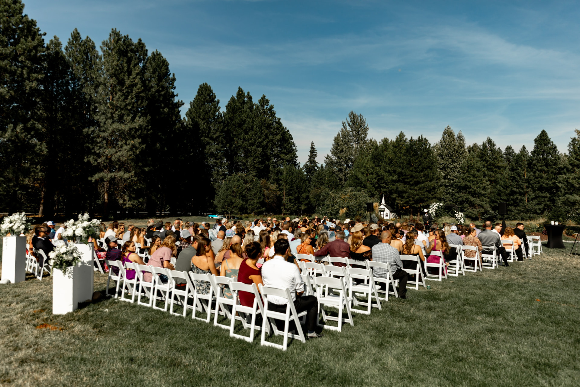 Rows of white resin folding chairs seating wedding ceremony guests on a lawn surrounded by pine trees