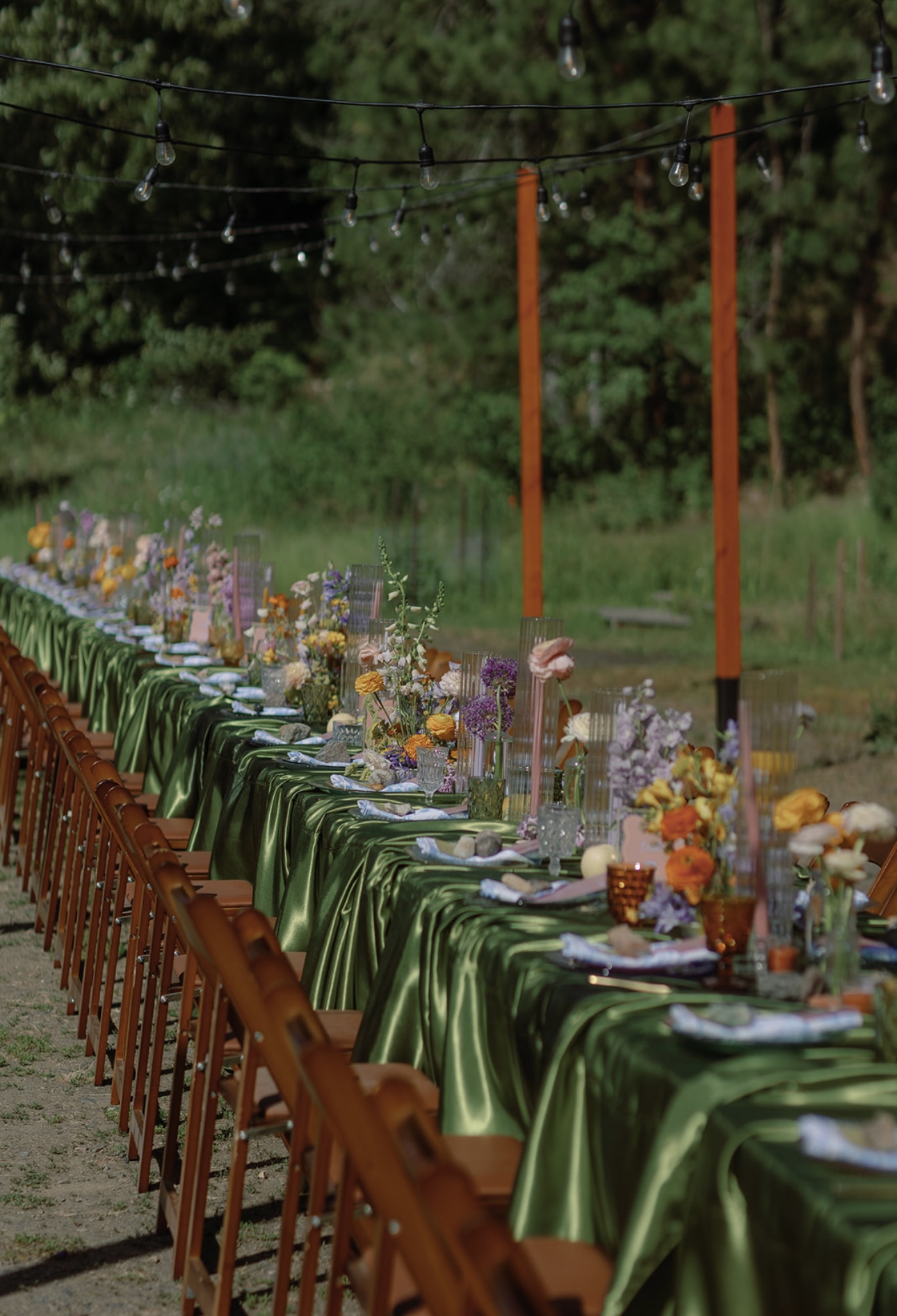 Long banquet table dressed in green satin linens, layered florals and glassware lit by overhead bistro string lights in a woodland reception setting