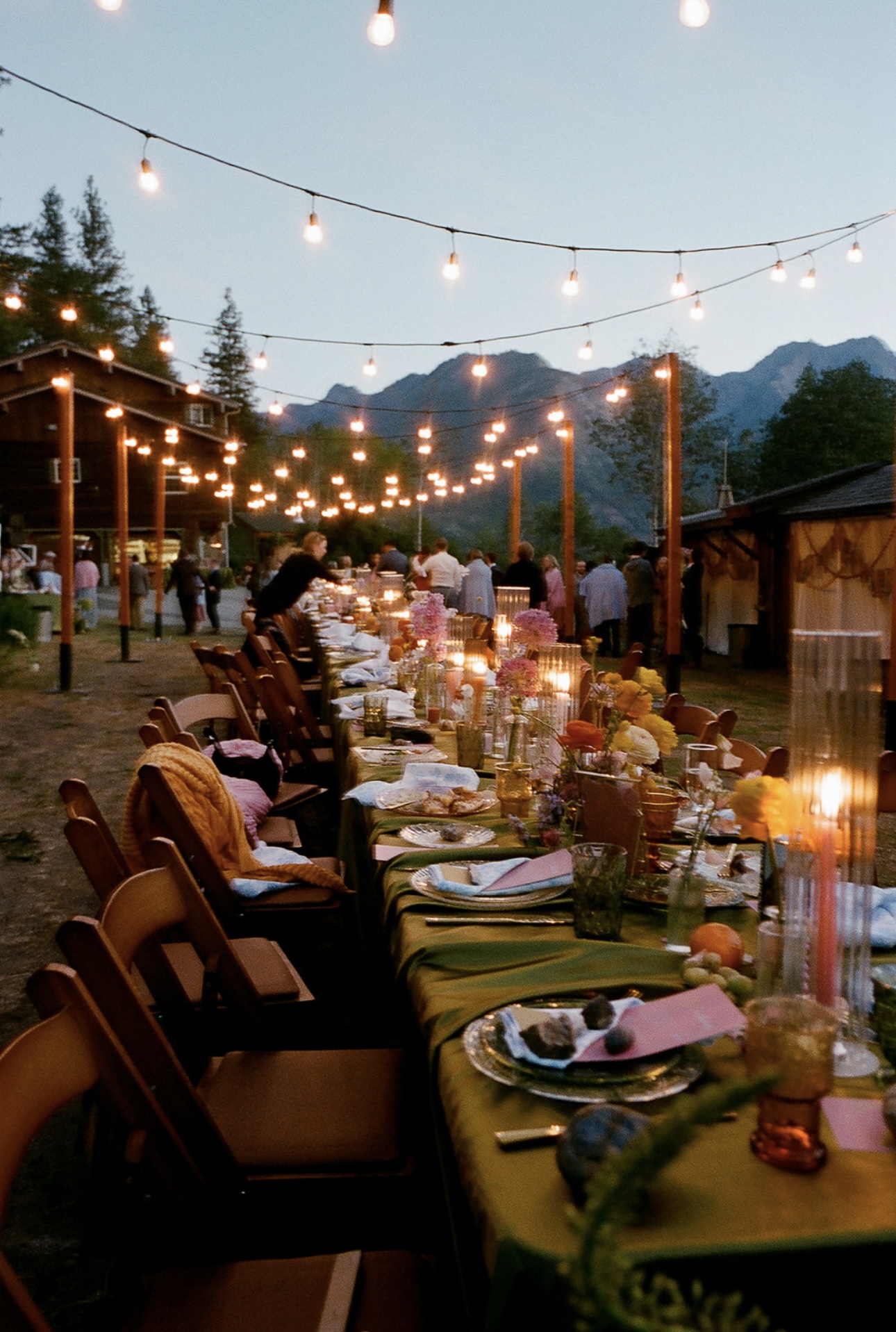 Warm bistro string lights glowing over a long family-style dinner table with green linens, colorful florals and candles at dusk, with Cascade mountains behind — outdoor event lighting by NCW Event Rentals in Eastern Washington