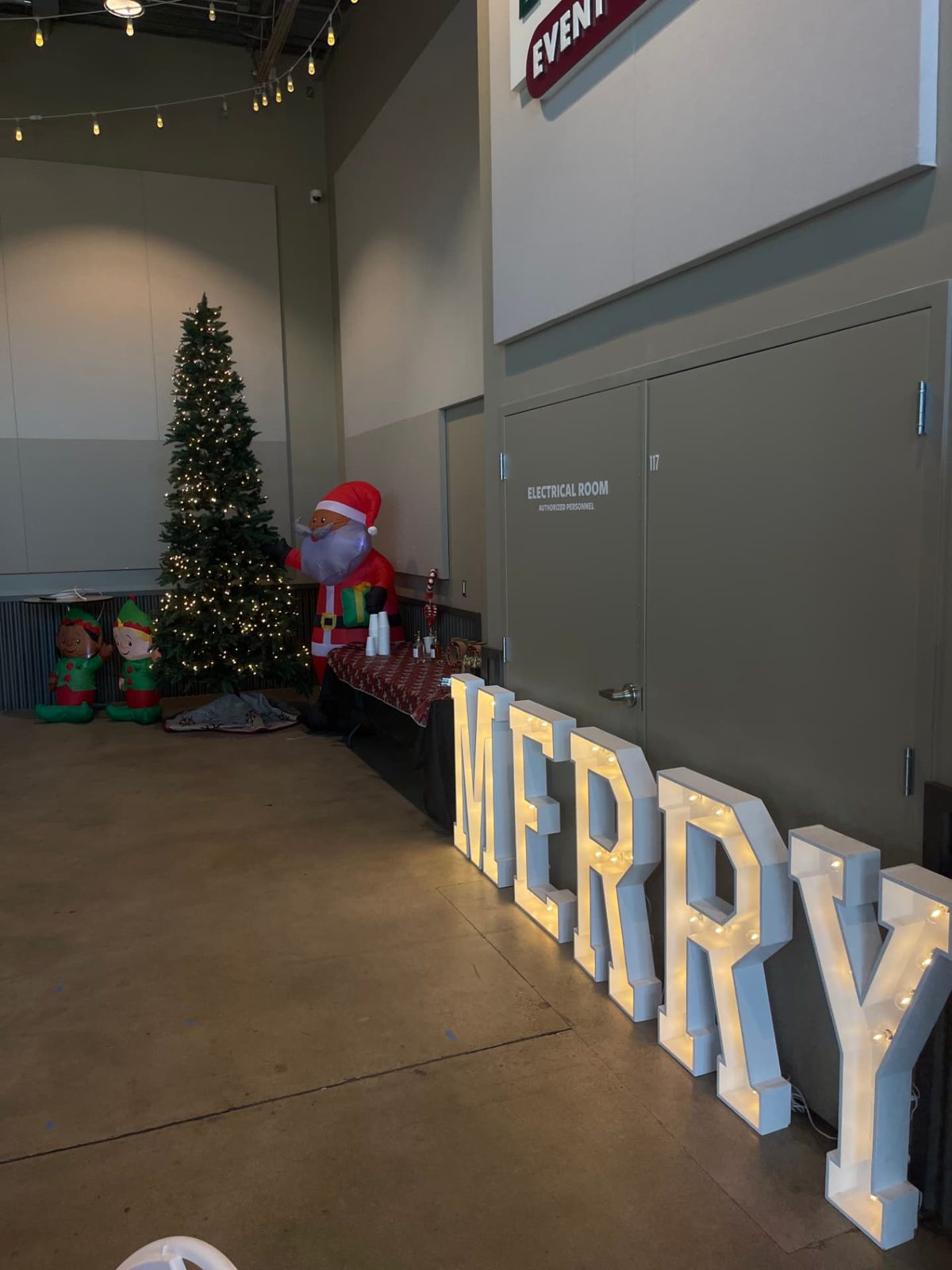 Lit MERRY marquee letters beside a decorated Christmas tree and inflatable Santa at a holiday event