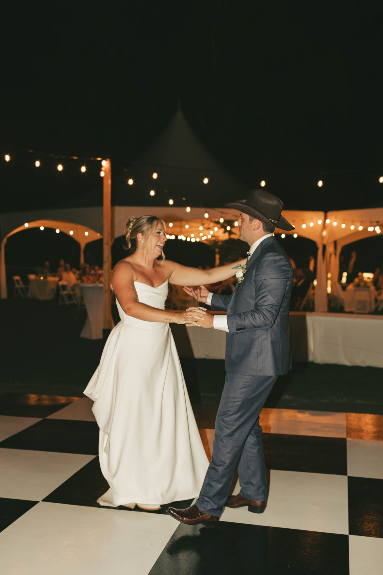 Newlyweds dancing on a black and white checkered dance floor under tent string lighting
