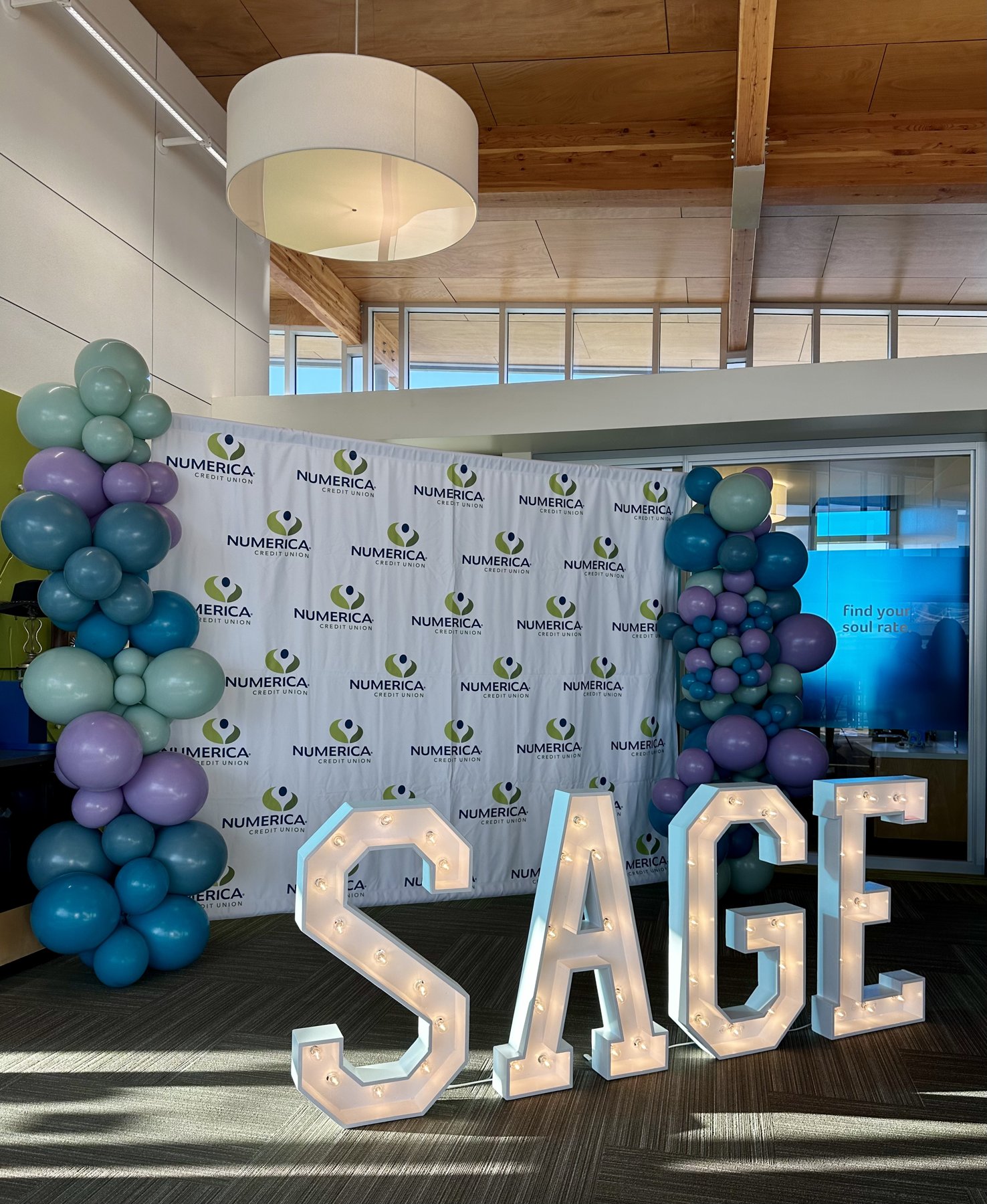 Lighted SAGE marquee letters in front of a Numerica Credit Union step-and-repeat backdrop with balloon columns at a corporate launch event