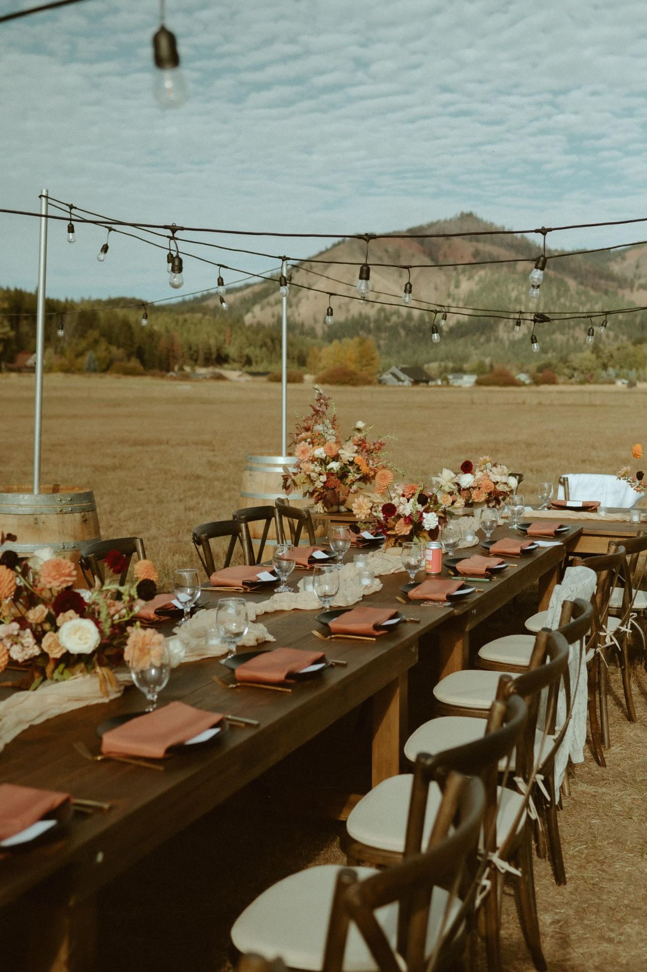 Wood farm tables with cross back chairs, terracotta napkins and wine barrels under string lights with a mountain view