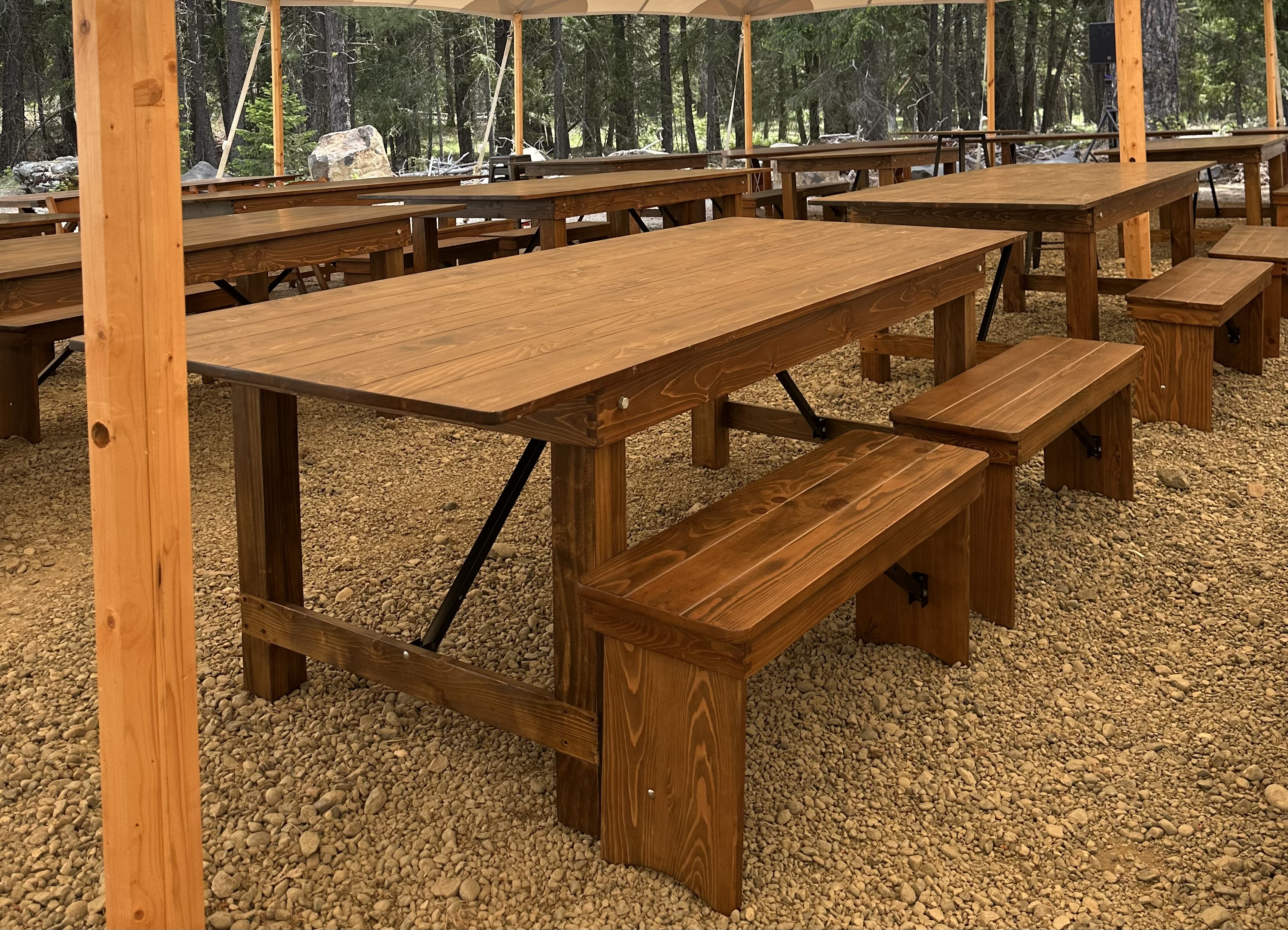 Rustic wooden farm tables and benches arranged under a white canopy tent on a gravel clearing in the Suncadia pine forest near Cle Elum, WA — corporate dining setup by NCW Event Rentals