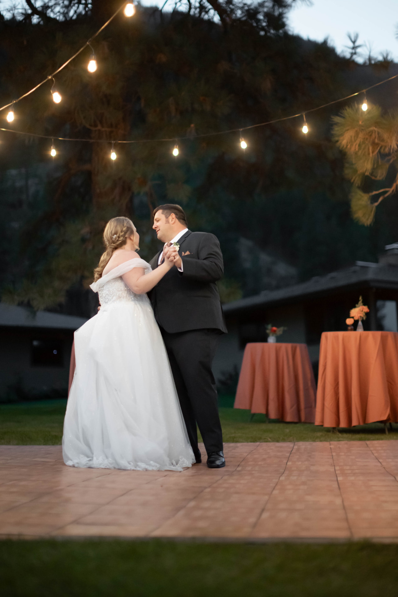 Bride and groom dancing under bistro string lights beside terracotta linen cocktail tables at an evening reception