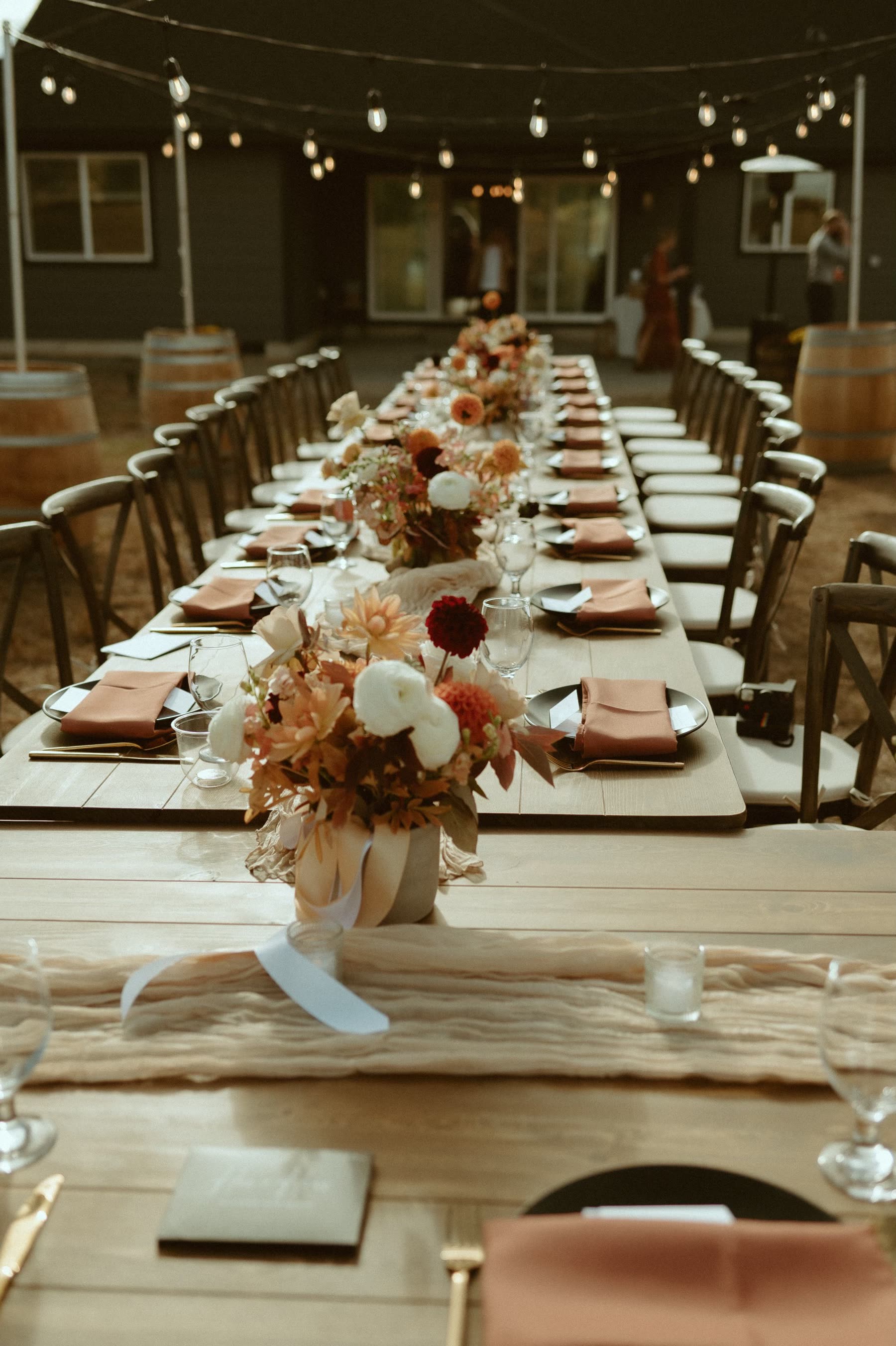 Long farmhouse banquet row with gold flatware rentals, terracotta napkins and matte black plates under bistro lighting at an Eastern Washington reception