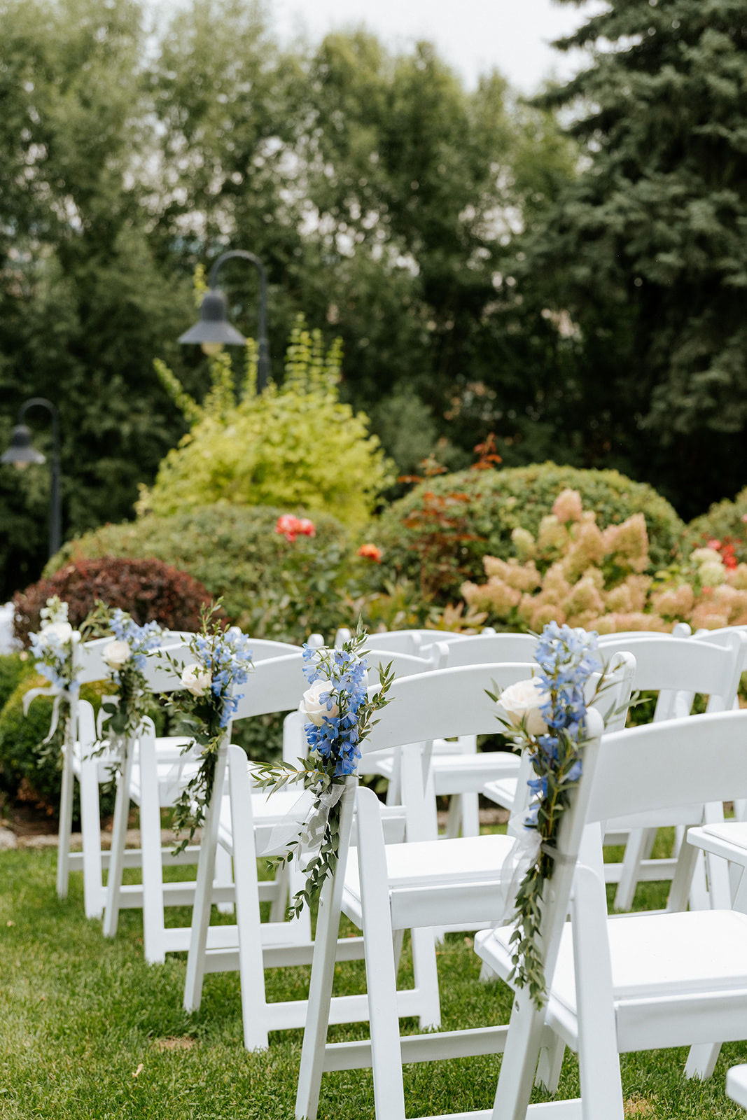 White resin folding chair rentals with blue delphinium and white rose aisle florals at an outdoor wedding ceremony