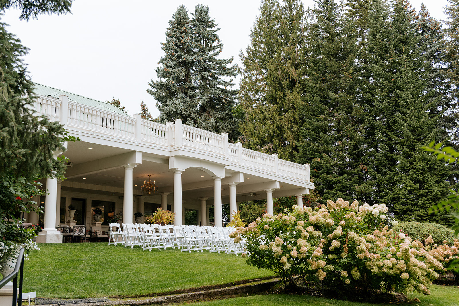 White folding chairs set for a ceremony on the lawn of a white-columned estate venue with hydrangeas