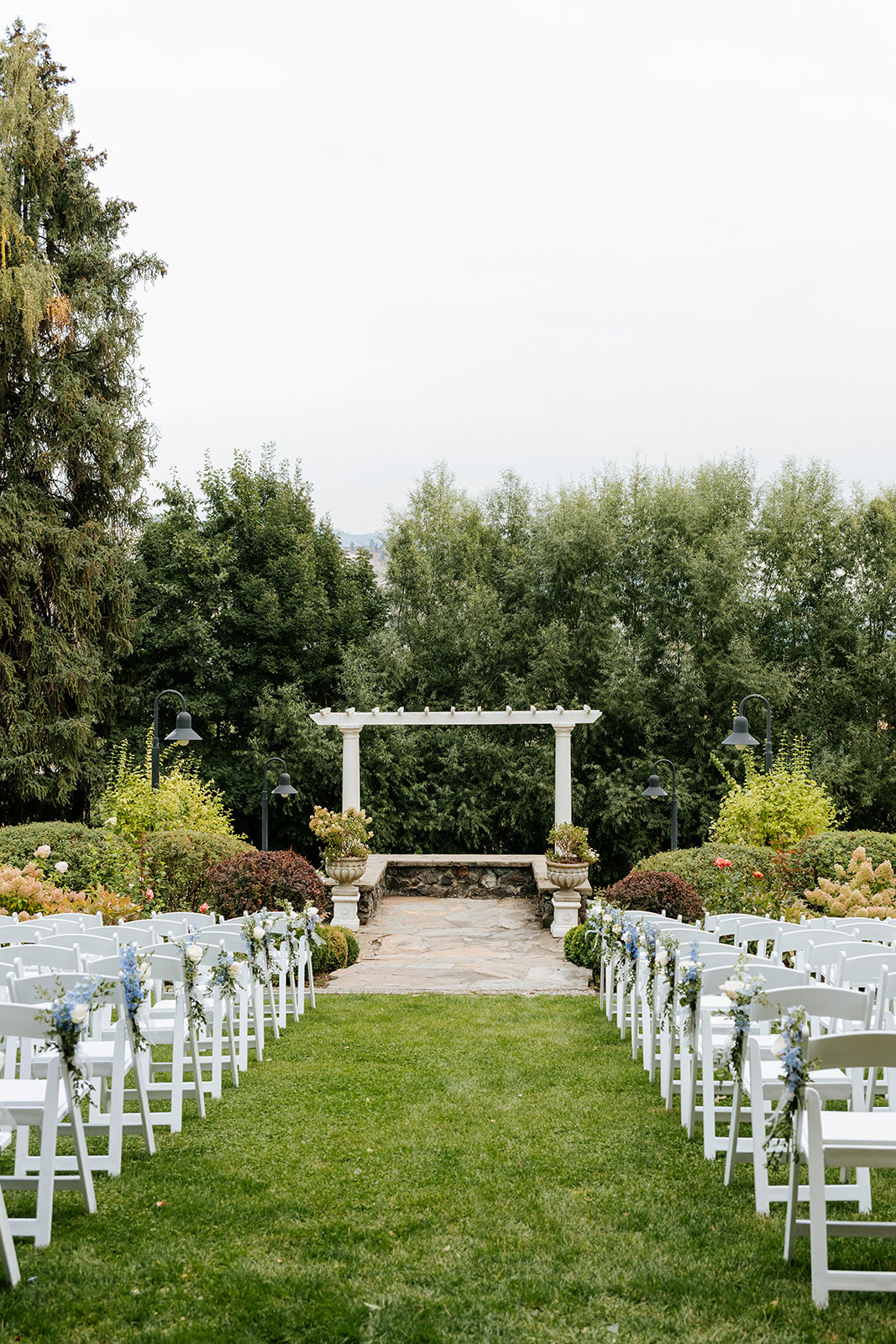 Wedding ceremony aisle at Beecher Hill House in Wenatchee lined with white folding chair rentals and blue delphinium florals facing the stone pergola altar