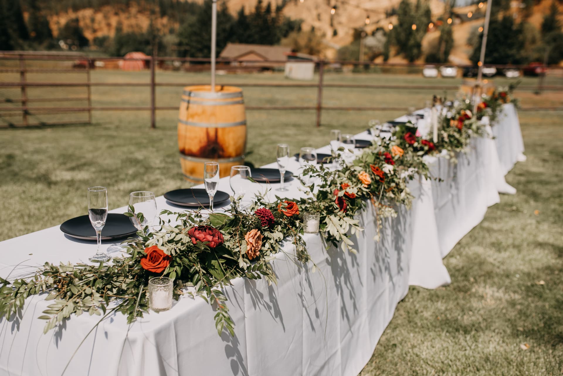 Long head table with white linen, black plates and floral garland beside a wine barrel at an outdoor ranch wedding