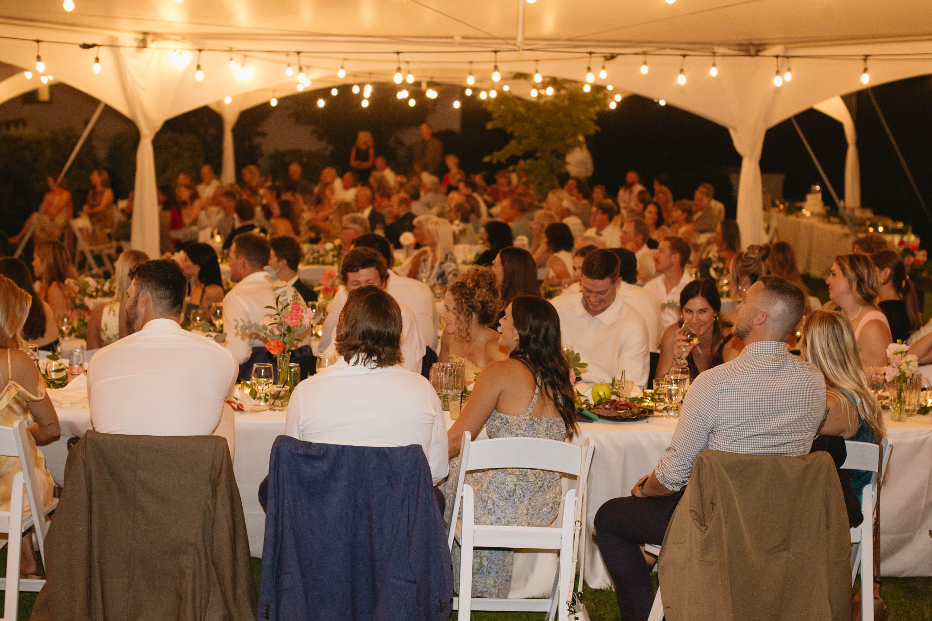 Guests seated at long banquet tables with white linens under a lit wedding tent with cafe string lights at night