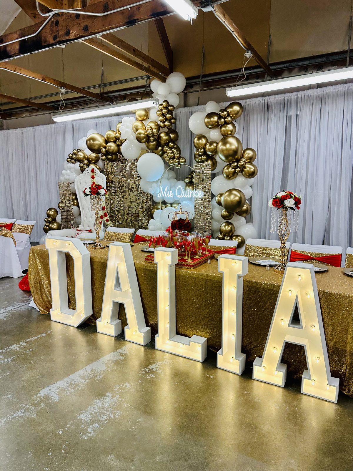 Lit marquee letters spelling DALIA in front of a quinceañera head table with a black and gold balloon garland