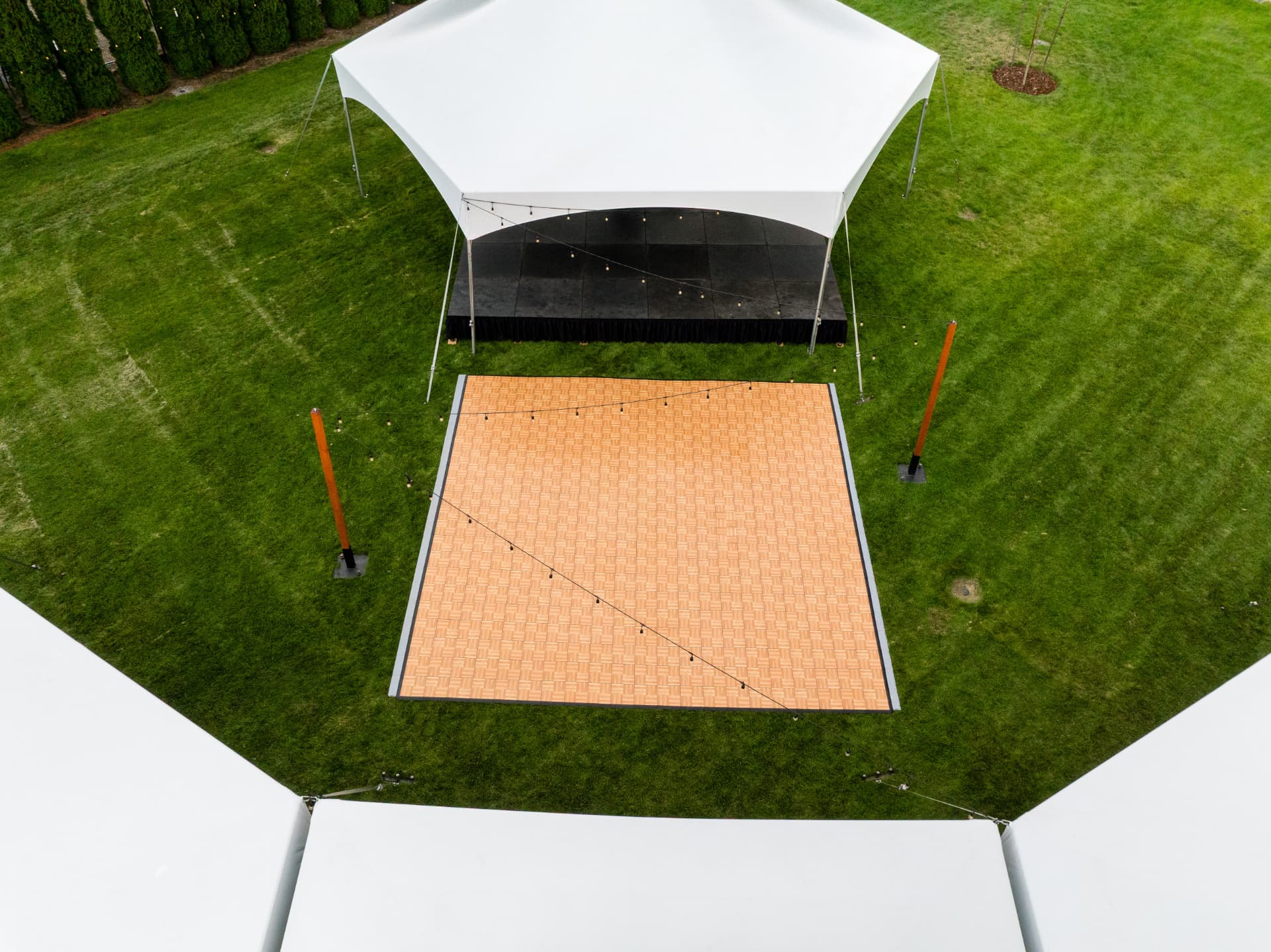 Aerial view of string lighting strung over a parquet dance floor and black stage between tents