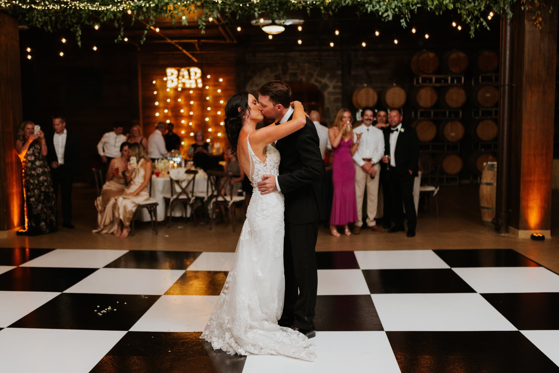 Bride and groom kissing on a black and white checkered dance floor in front of a lit BAR marquee at a winery wedding reception