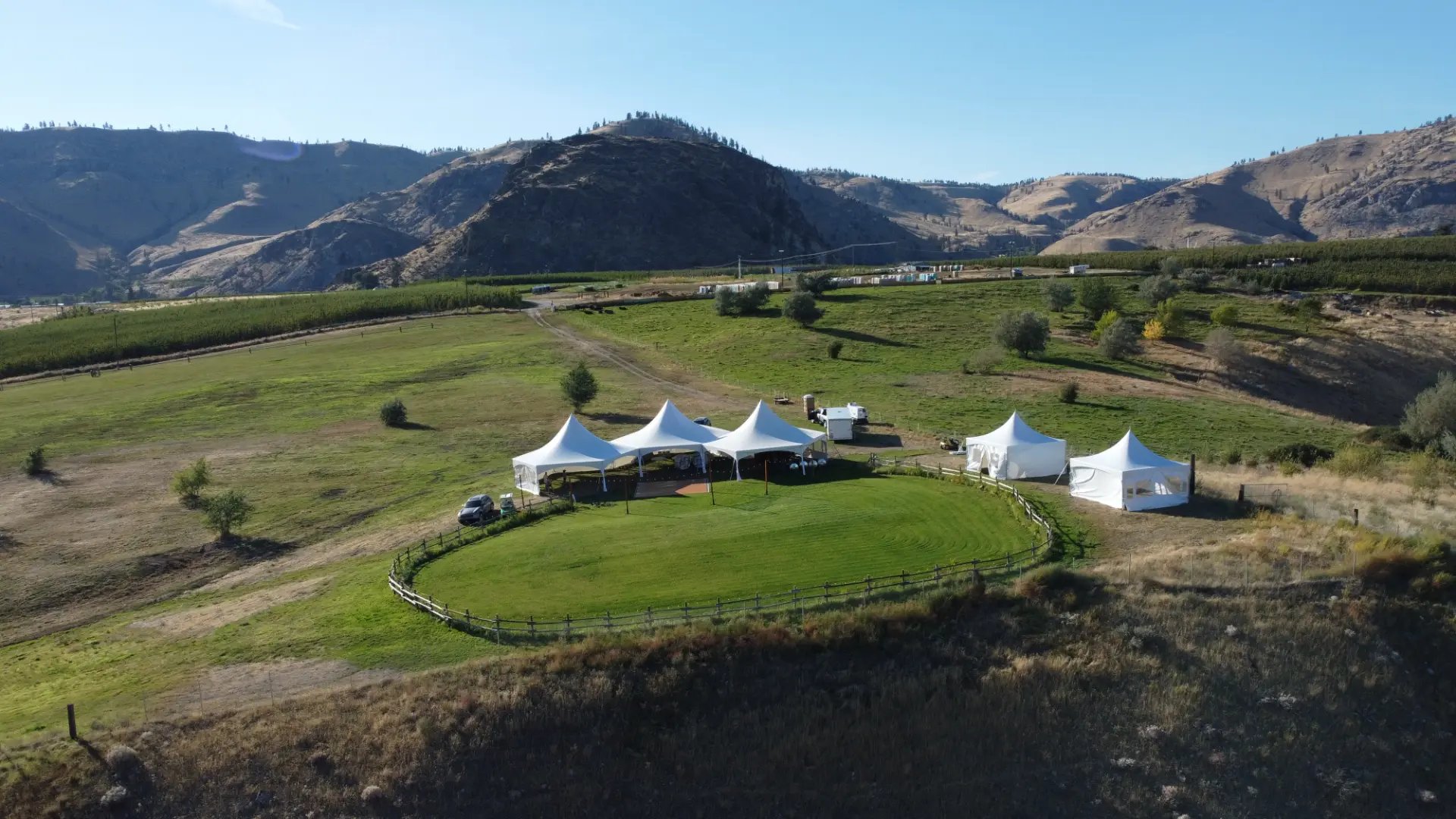 Wide aerial view of a Chelan private property wedding with the three connected hexagon tents of the 3 Hex Tent Crescent Package set on a circular lawn, mountains beyond — tent rental from NCW Event Rentals