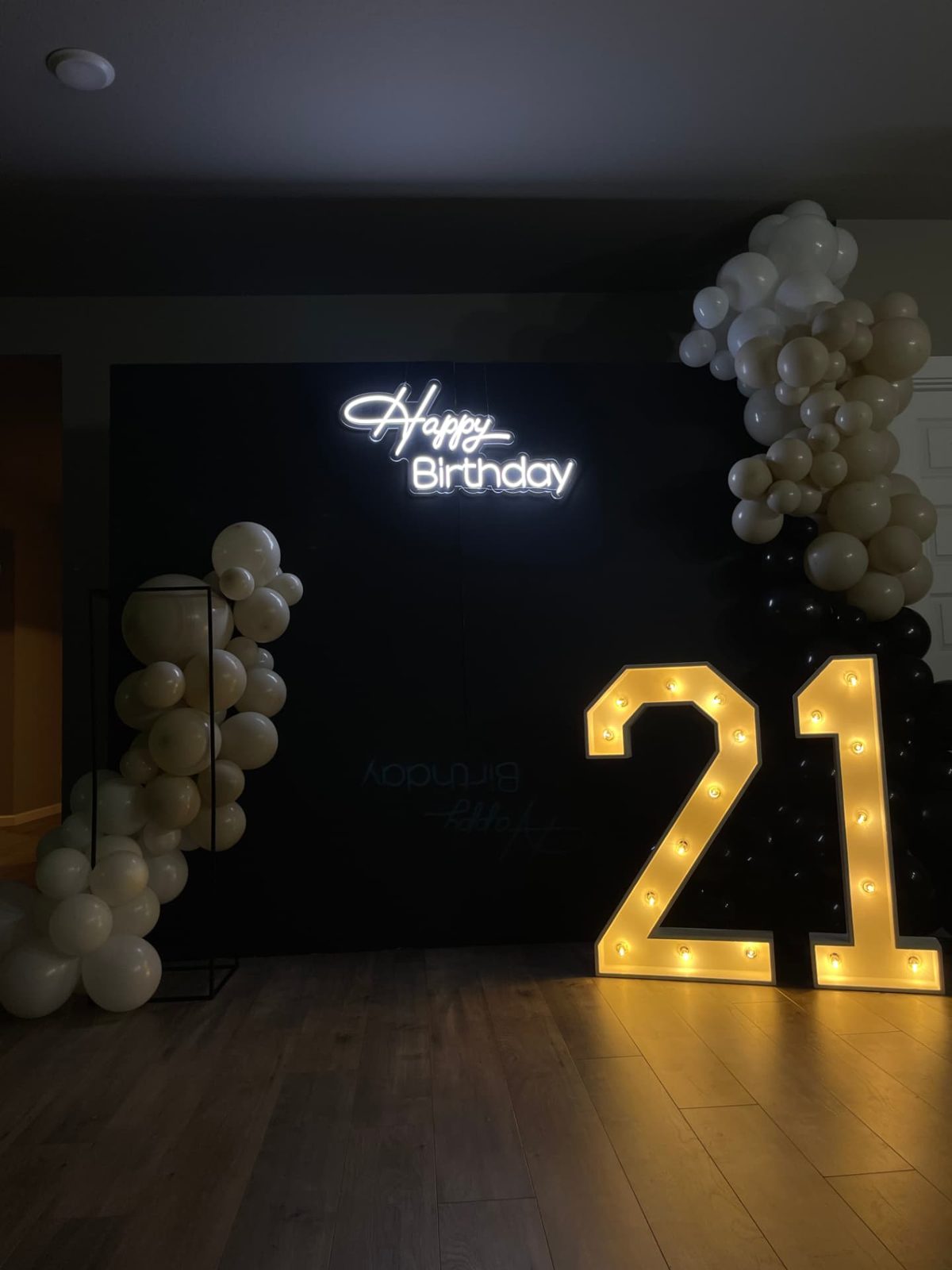 Lit 21 marquee numbers beside a black backdrop with a Happy Birthday neon sign and cream balloon garlands