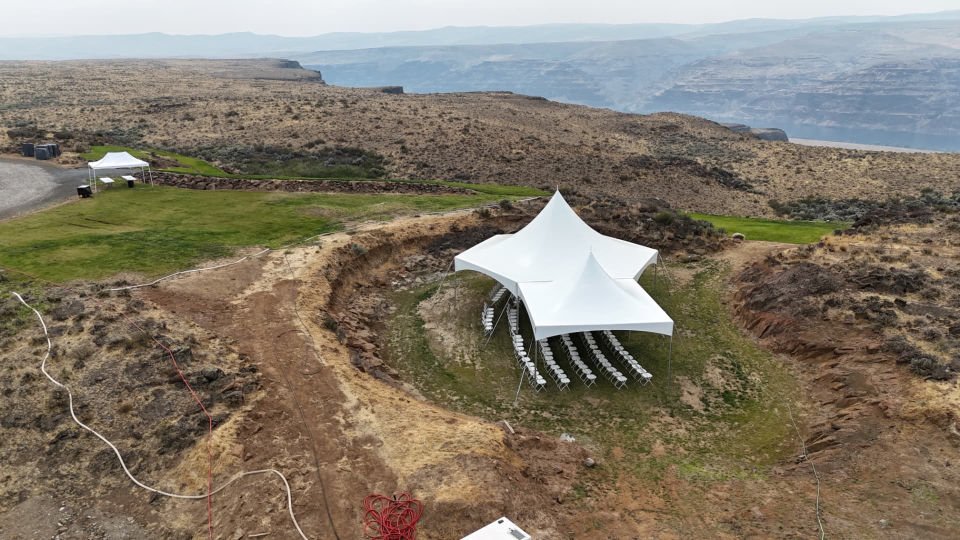 Aerial view of a white high-peak ceremony tent set into a canyon overlook with rows of folding chairs facing the river