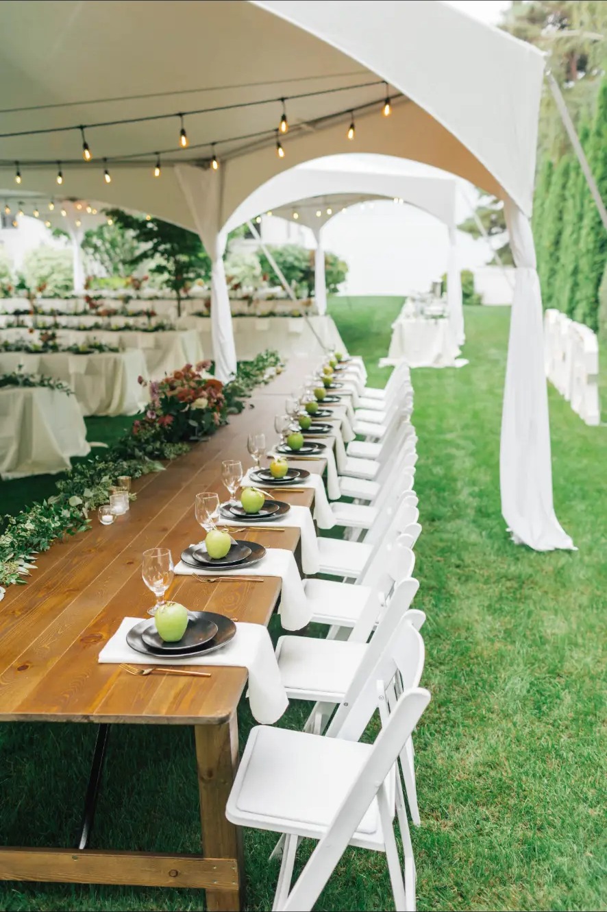 White folding chairs lined along a long farmhouse table under a tent with bistro string lights