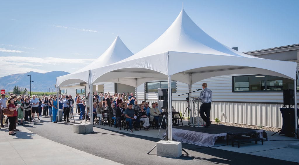 Crowd seated under twin white high-peak tents facing a speaker on a black stage deck at a corporate facility opening