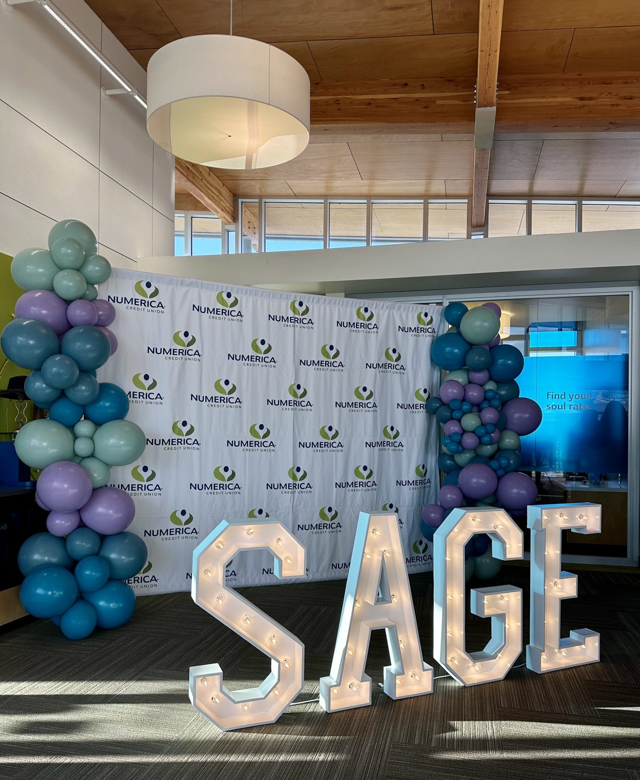 SAGE marquee letters lit in front of a Numerica Credit Union step-and-repeat banner with balloon columns