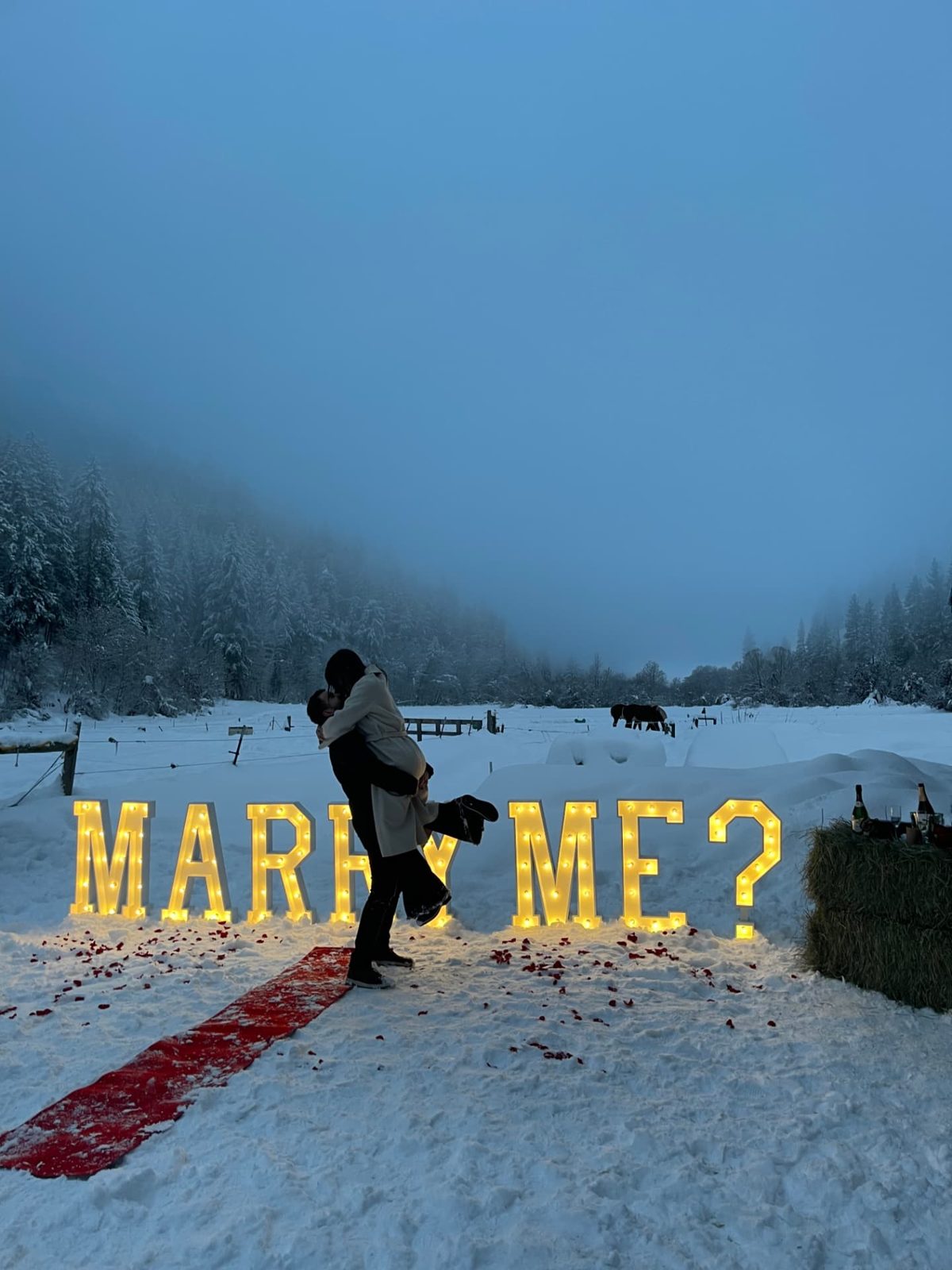Lit MARRY ME? marquee letters in fresh snow with a red carpet and rose petals at a winter proposal near Leavenworth, Washington