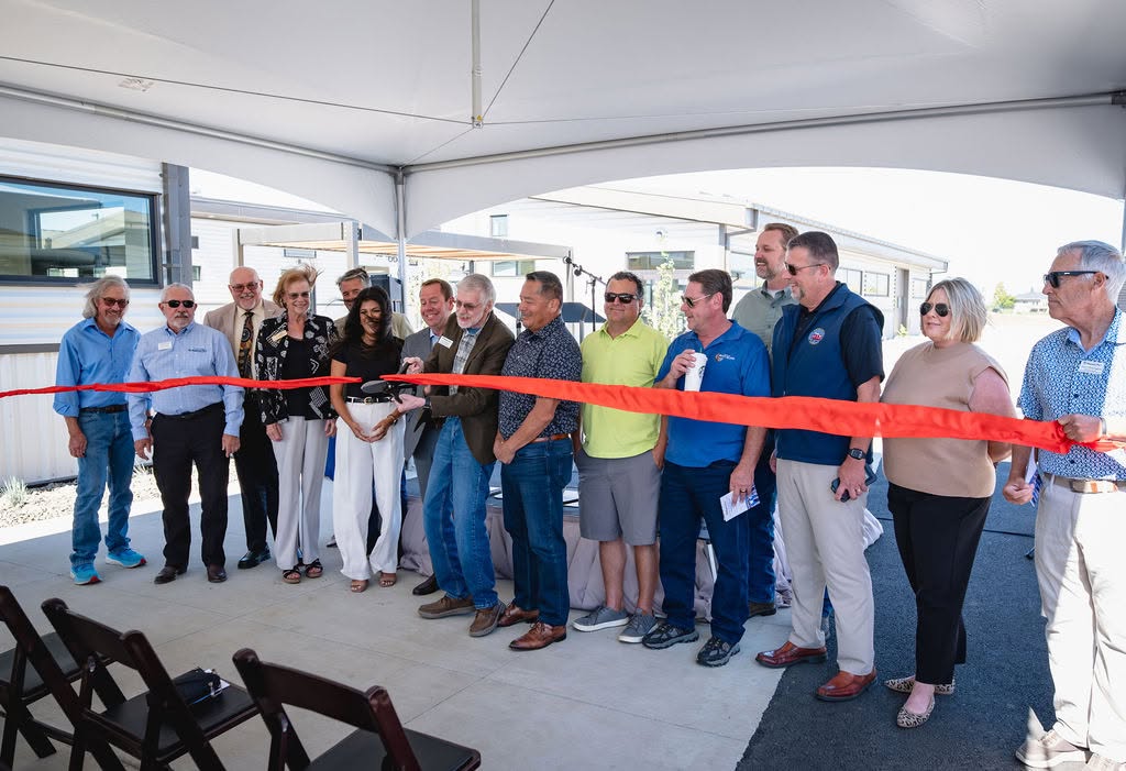 Officials cutting a red ribbon under a white high-peak tent with padded folding chairs at a corporate grand opening
