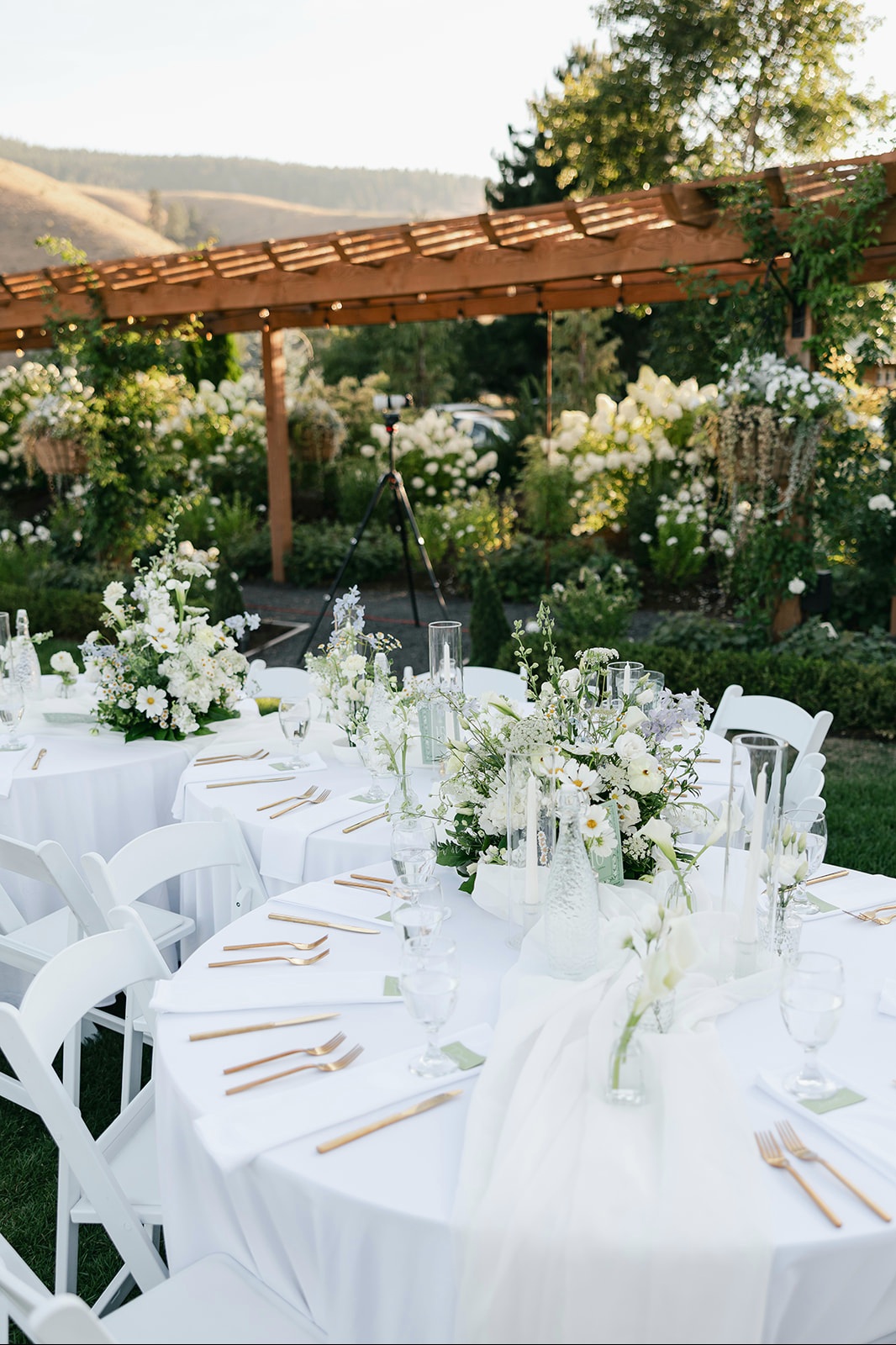 Round table rentals with white linens, white folding chairs and white floral centerpieces under a timber pergola at a garden wedding reception