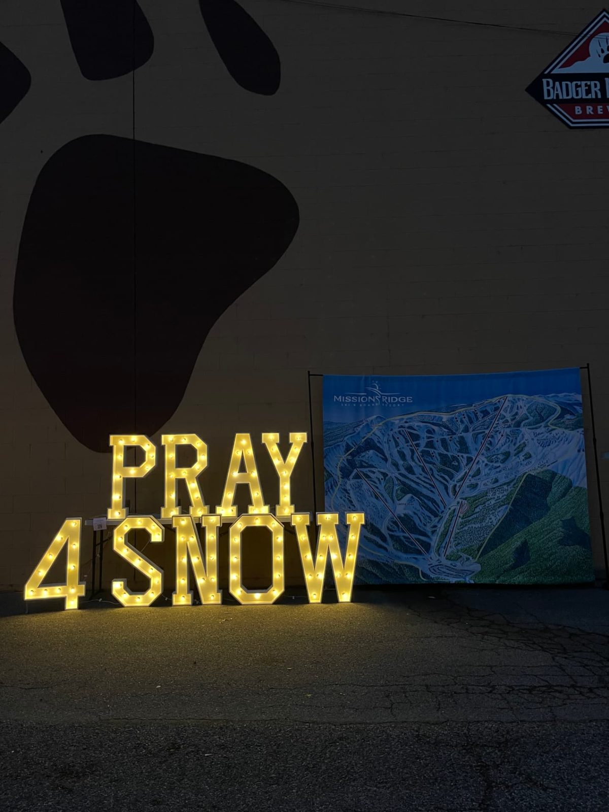 Lit PRAY 4 SNOW marquee letters next to a Mission Ridge trail map banner at a ski season kickoff event in Wenatchee