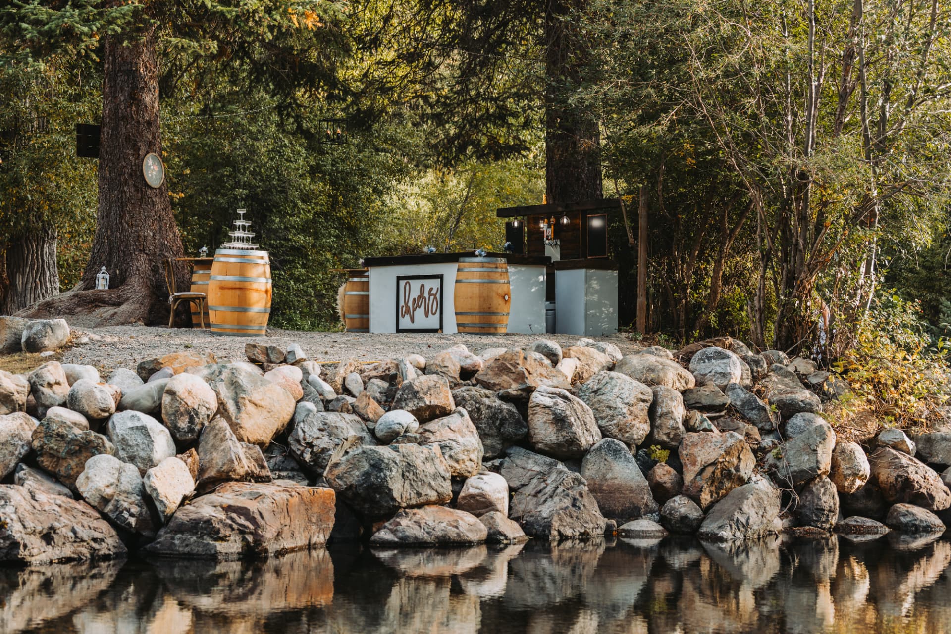 Rustic outdoor bar with white paneled front, wine barrels and a champagne tower set in a forest clearing beside a rocky lake shore near Suncadia Resort in Cle Elum, WA