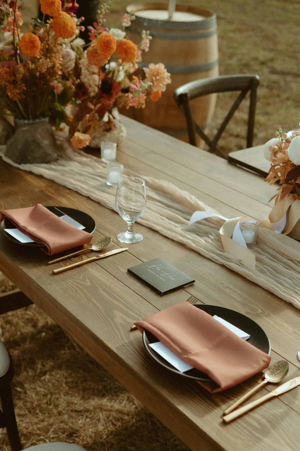 Wood farm table place setting with black plates, gold flatware, terracotta napkins and a gauze runner at a tented wedding