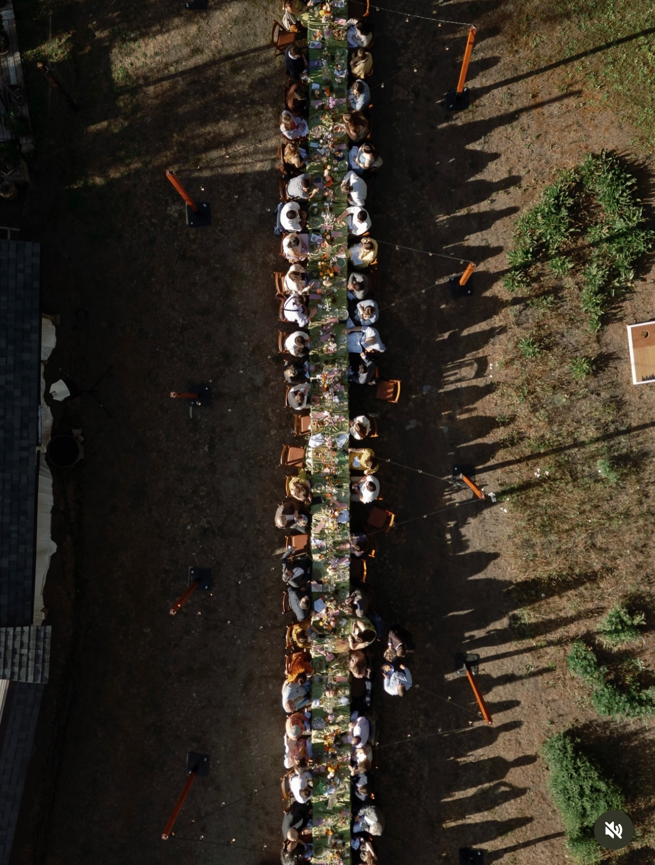Aerial view of a long family-style dinner table lined with bistro string lights, guests seated on both sides, next to an orange barn with a white star painted on the roof