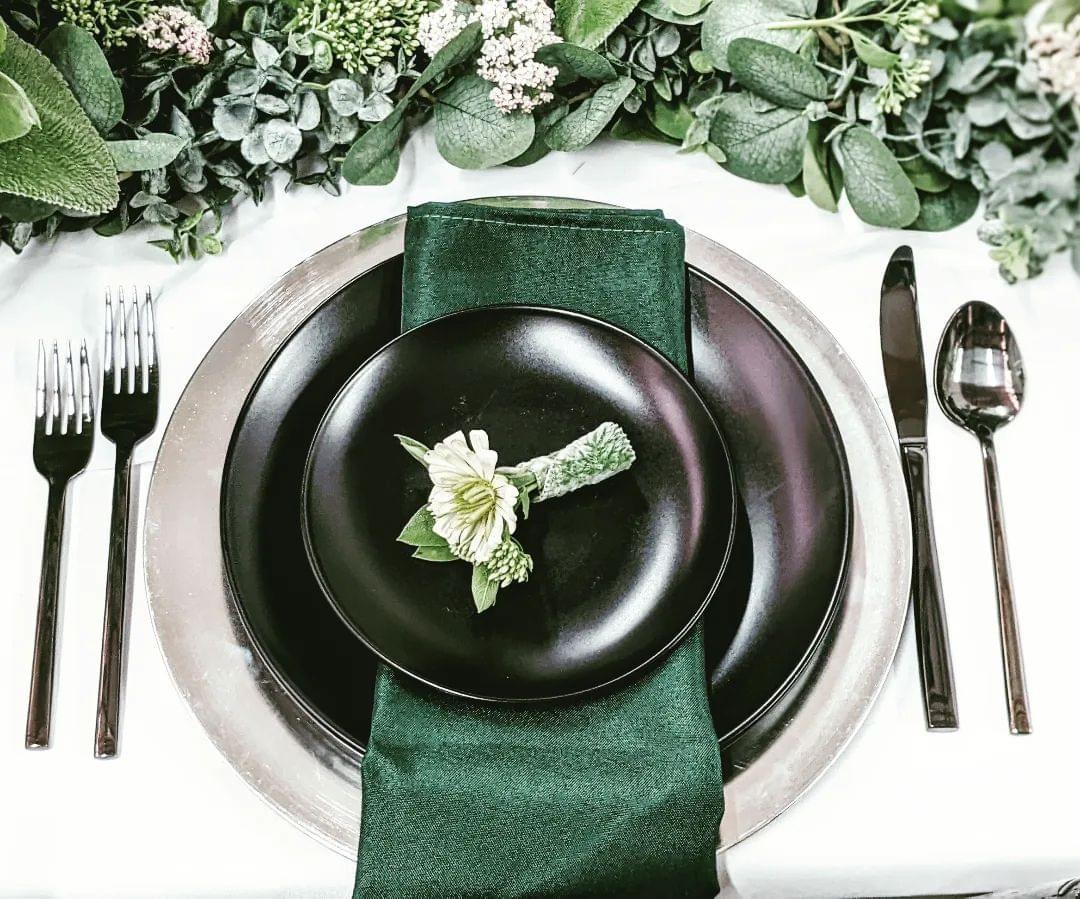 Place setting with silver charger plate, matte black plates, emerald green napkin and polished flatware rentals