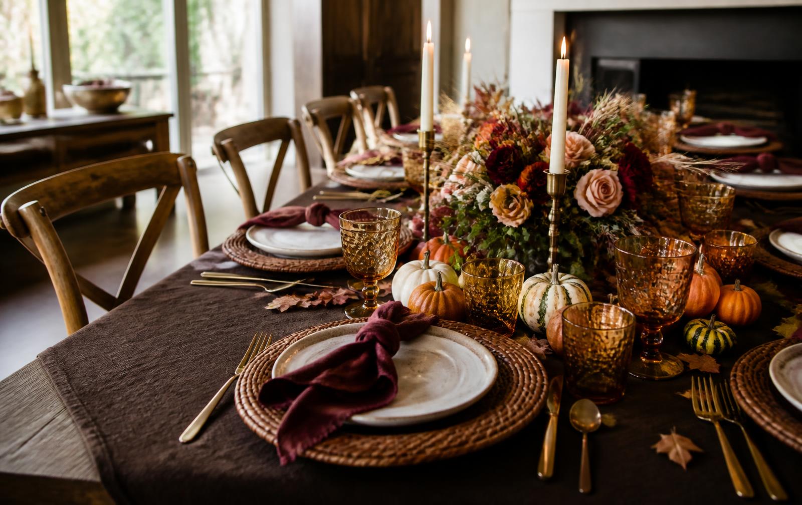 Thanksgiving dinner table set with brown linen, burgundy napkins, wicker chargers, amber glassware, candles and an autumn floral centerpiece
